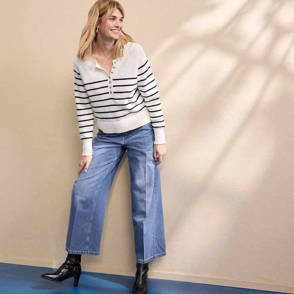 Pruhovaný bílý svetr na zip v kombinaci se širokými džínami a černými kozačkami.