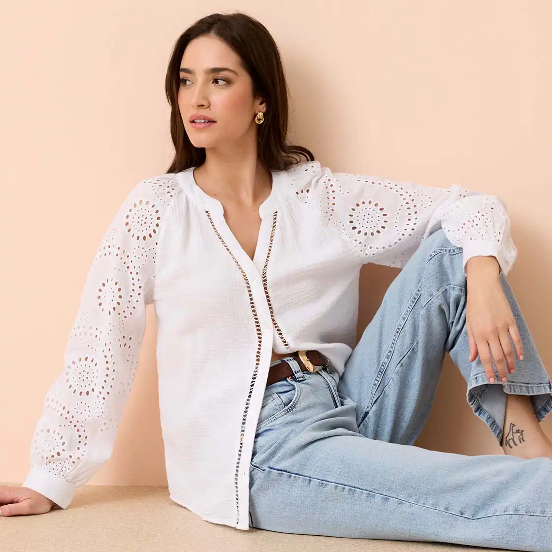 Bílá halenka s dírkovanou výšivkou a dlouhým rukávem, v kombinaci se světle modrými džínami a opaskem.