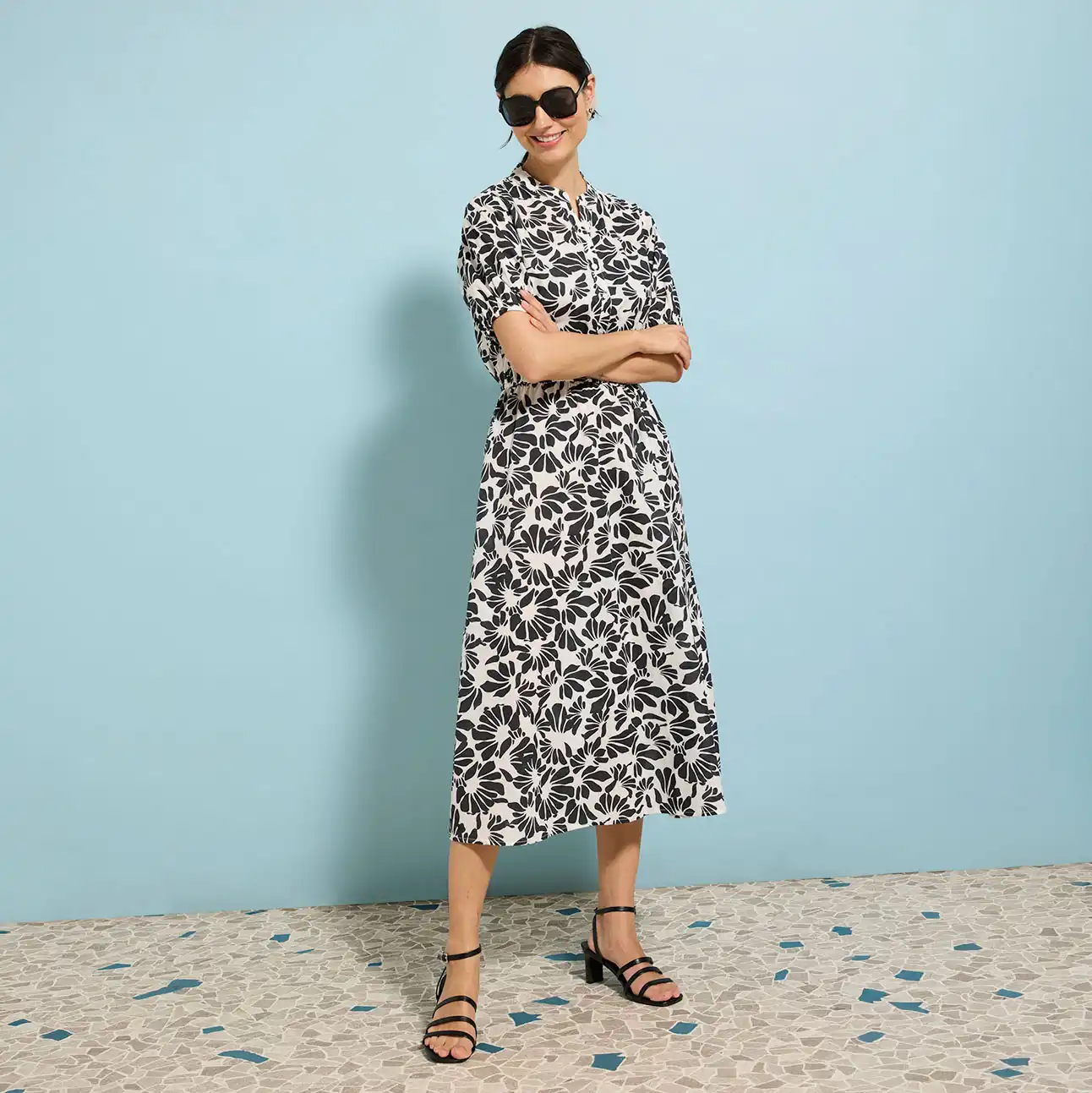 Letní midi šaty s černo-bílým květinovým vzorem, sluneční brýle a ploché sandály vytvářejí elegantní outfit.