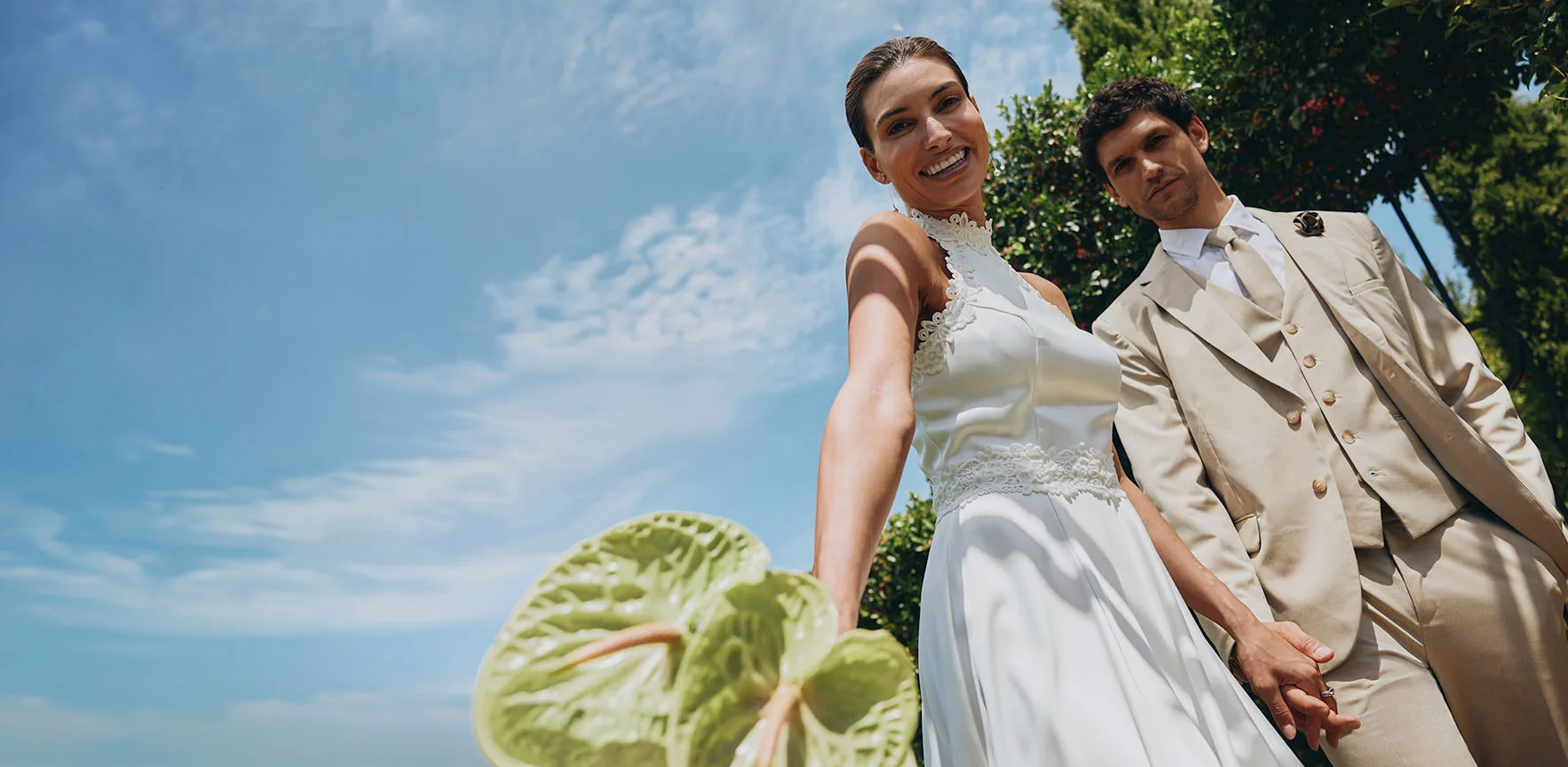 Elegantní novomanželský pár v záři slunce: ona v bílých šatech, on v béžovém obleku a se slunečními brýlemi.