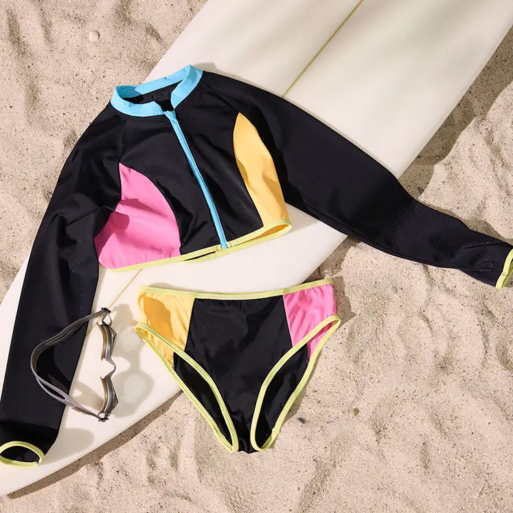Barevné plavky s dlouhým rukávem a bikiny na písku vedle surfovacího prkna.