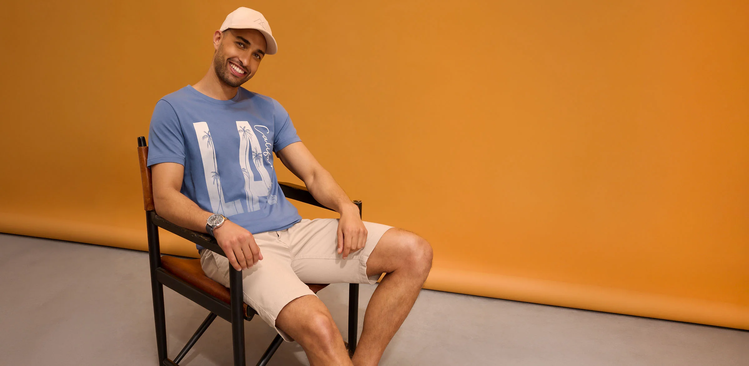 Ležérní outfit: kšiltovka, modré triko s potiskem, béžové šortky a náramkové hodinky.