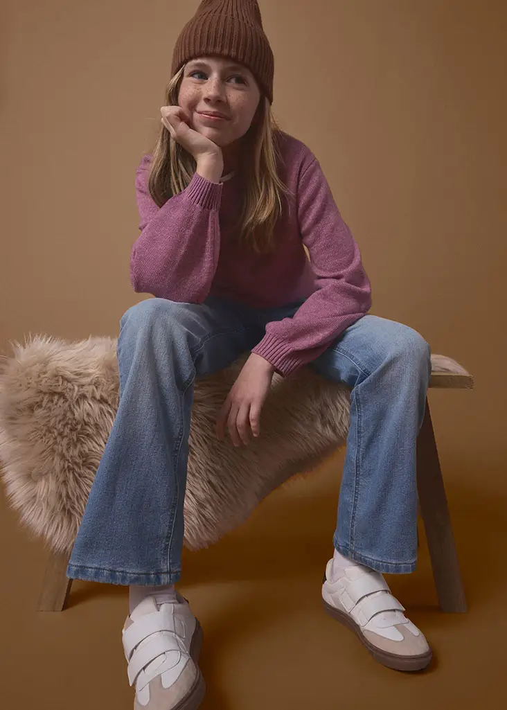 Dívka má na sobě hnědou čepici beanie, svetr v barvě pink, modré džíny a bílé tenisky.